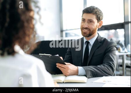 Successful caucasian man, hiring manager in a company, conducts an interview with a girl, in a modern office, looks through the candidate's resume, makes notes, asks questions, makes a decision Stock Photo