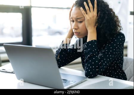 Exhausted African American woman, auditor, lawyer, economist, sits in front of a computer at a work desk in a modern office, holds her head, cannot cope with a hard work task, needs help and rest Stock Photo