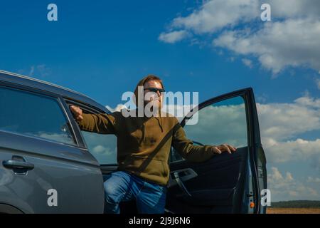 Young man weared in hoody and sun glasses holding his luxury car door and looking far away. Stock Photo