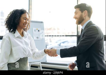 Successful influential business colleagues, young adult Caucasian man and African American woman, completed business negotiations, made a good deal, signed a contract, greet each other, shake hands Stock Photo