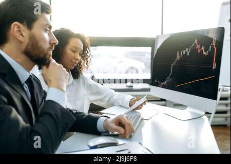 Successful colleagues traders, a Caucasian man and an African American woman, sit in front of a computer screen, work with trading charts, analyze the cryptocurrency market, predict the dynamics Stock Photo