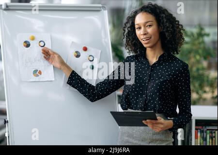 Intelligent young adult African American woman, financial analyst, economist, stands in the office near the whiteboard, presents the project, demonstrates financial charts, looks at camera, smiling Stock Photo