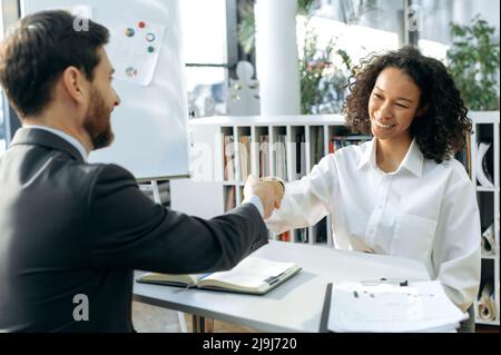 Smart successful African American woman was hired by the company. Two multiracial business colleagues, agreed to cooperate on a project, shake hands while sitting at a table in a modern office, smile Stock Photo