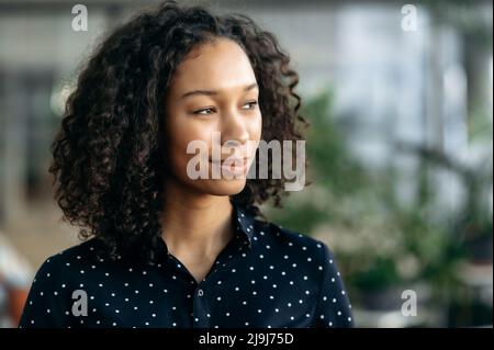 Close-up photo of a pretty, pensive dark-skinned girl, student, manager, secretary, freelancer or company manager, standing in the office, wearing stylish business clothes, looking away, smiling Stock Photo