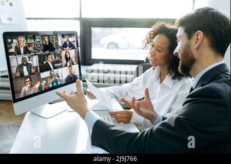 Business colleagues, co-founders of the company, sit at a table in the modern office in front of a computer screen, hold an online video conference with international colleagues, brainstorming Stock Photo
