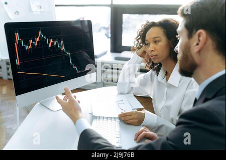 Multiracial business colleagues, invest in the stock market, co-founders of the company, sitting in front of a computer screen, analyze the dynamics of quotes charts, predict the dynamics Stock Photo