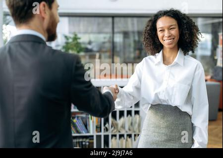 Making a deal, shaking hands, confirms agreements. African american successful business woman, sales manager, shakes hands with male colleague or client in modern office, smiling at each other Stock Photo