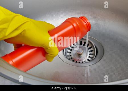 Person's hand in a yellow rubber glove pours pipe cleaner down the drain of a metal kitchen sink Stock Photo