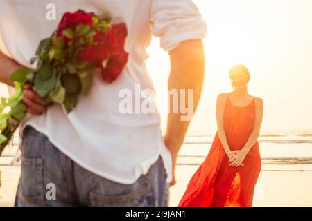 happy beautiful woman in red dress receiving a gift from a man by sea sunset.surprise wife from husband bouquet of roses women's day March 8 and Stock Photo