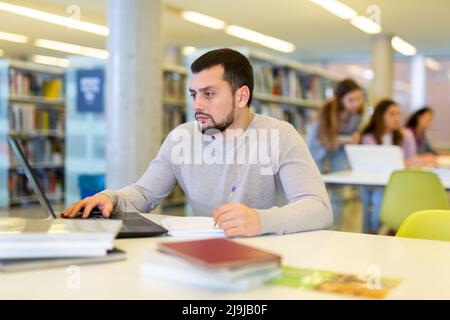 Focused young student studying with books and laptop in library Stock Photo