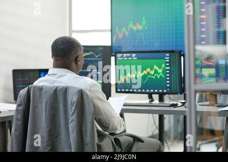 Rear view of unrecognizable African American man sitting in front of computer monitors wokring with currency stats Stock Photo