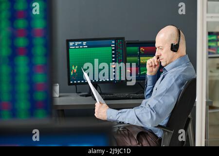 Mature professional Caucasian stock data analyst sitting at desk in office working with financial documents Stock Photo
