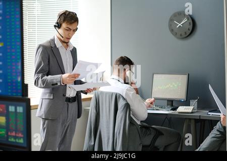 Two men in formalwear working in brokerage company having phone calls with their business partners and clients and looking through documents and stats Stock Photo