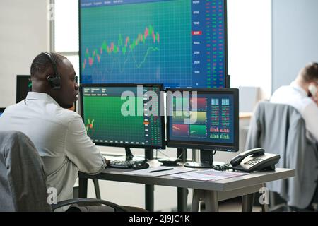 African American and Caucasian men in formalwear sitting in front of computer monitors analyzing currency stats Stock Photo