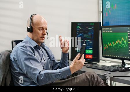 Mature Caucasian man wearing headset with mic sitting in office speaking to investor on video call using smartphone Stock Photo