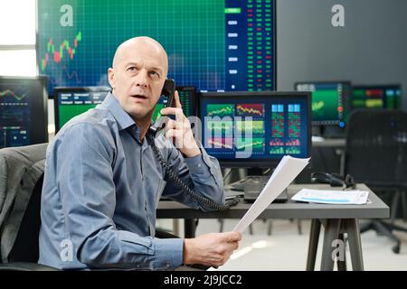 Portrait of mature stock trading specialist holding documents looking at monitor with stock trading results and talking on phone Stock Photo