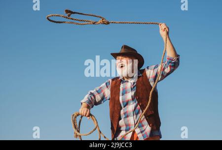 Old wild west cowboy with rope. Bearded western man throwing lasso with brown jacket and hat catching horse or cow. Stock Photo