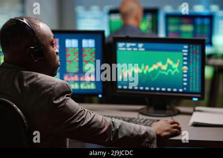 Rear view of African American man sitting in front of computer monitors watching and analyzing trading stats in evening Stock Photo