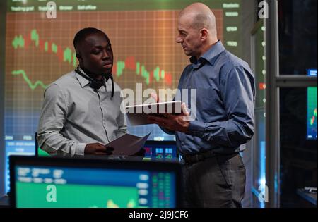 Two ethnically diverse men working in stock trading and investment company looking at charts and graphs on digital tablet and discussing them Stock Photo