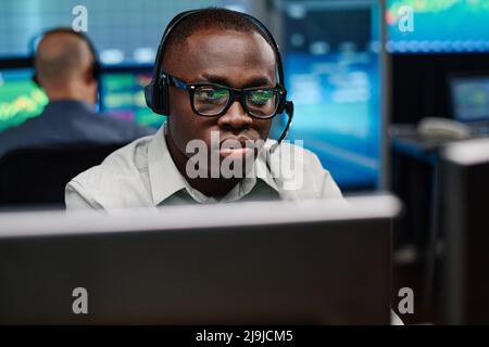 Serious young African American man wearing eyeglasses working in stock trading company watching charts and graphs of currency on computer screen Stock Photo