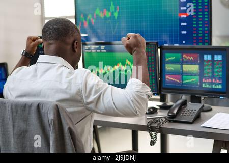 Rear view of young Black man in formalwear looking at computer screen watching stock trading graph growth and feeling excited Stock Photo
