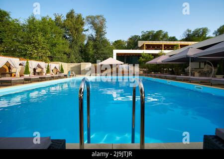 Swimming pool area with ladder, gazebos, deck chairs and sun umbrellas along the poolside in luxury resort. Summer houses surrounded with greenery Stock Photo
