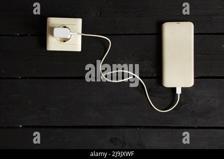 a white phone in a case is on charging in a socket on a black wooden background, the phone is on recharging Stock Photo