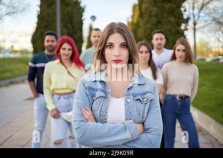 Self assured young female with crossed arms in denim jacket looking at camera while standing on street against friends on city park Stock Photo