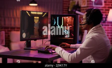 Handheld shot of african american man using pc gaming setup relaxing playing first person shooter in multiplayer. Gamer streaming while explaining gameplay in online action game talking to team. Stock Photo