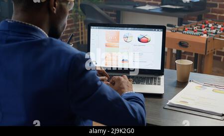 Office employee working with data statistics at desk, using laptop to plan business charts and browsing internet. Project manager analyzing diagrams and paperwork on online network. Stock Photo