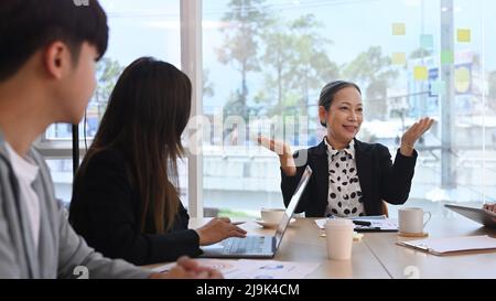 Concentrated middle aged female team leader explaining project details to young employees at meeting Stock Photo