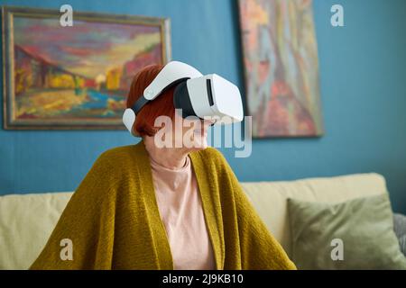 Grandmother in virtual reality goggles playing online while resting on sofa in room Stock Photo