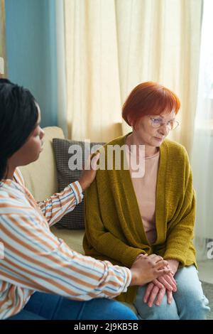 Depressed senior woman sitting on sofa with sad expression while caregiver talking to her and supporting her Stock Photo