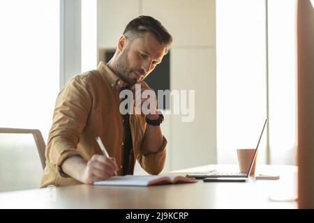 Thoughtful Businessman Writing Taking Notes Using Laptop At Workplace Stock Photo