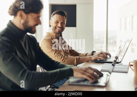 Happy male colleagues using laptops and communicating, working on business project together, discussing ideas in office Stock Photo