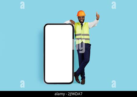 Smiling Black Construction Engineer Standing Near Blank Smartphone And Showing Thumb Up Stock Photo