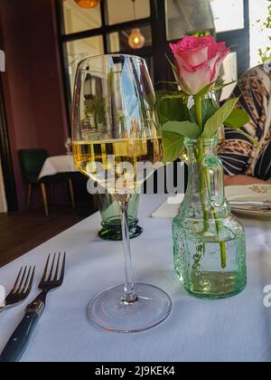 Glass of white wine with a pink rose on a table in a bistro with an unrecognizable woman in the background Stock Photo