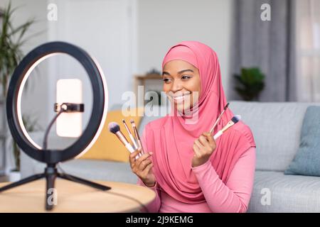 Muslim beauty blogger. Happy african american woman showing her brushes and doing makeup, recording vlog on cellphone Stock Photo