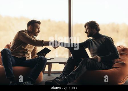 Male colleagues using computers, shaking hands, making business deal or agreement, sitting in bean bag chairs at office Stock Photo