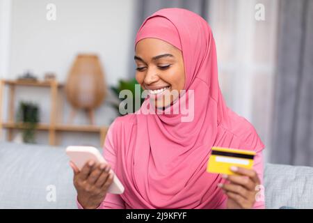 Happy african american muslim lady shopping online, using smartphone and credit card, purchasing from home Stock Photo