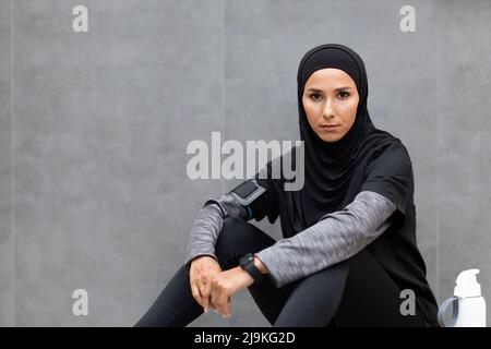 Serious confident young islamic lady in sportswear and hijab with bottle of water sits on floor