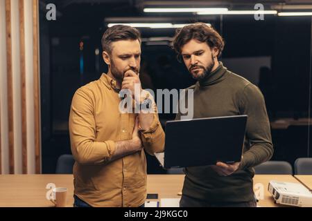Male colleagues looking at laptop screen, working on business project as team, staying at company office late in evening Stock Photo