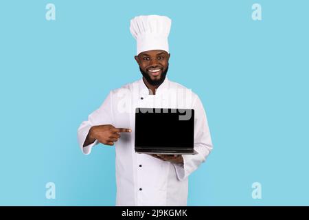 Cooking Website. Smiling Black Chef Pointing At Blank Laptop In His Hand Stock Photo