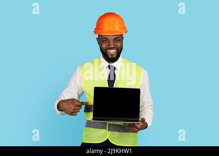 Smiling Black Construction Worker Pointing At Laptop With Blank Screen In Hand Stock Photo