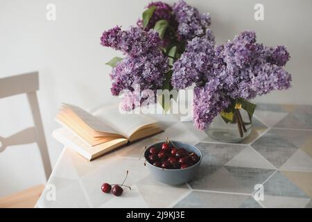 Romantic still life with an open book, lilac flowers and cherries on a table  Stock Photo