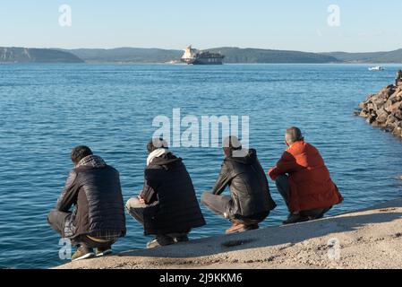 Turkish men squatting beside the Dardanelles Strait opposite the Gallipoli Peninsular, the harbour of the port city of Canakkale, Western Turkey. The Gallipoli Peninsula on the north bank of the Dardanelles strait in north-western Turkey, is the site of extensive First World War battlefields and memorials, a campaign that took place between the Ottoman Empire and the Allied powers. The Dardanelles Strait is an important commercial shipping route that connects the Mediterranean and the Black Sea, with the city of Canakkale at the entrance, a city that is not only important for its historical an Stock Photo