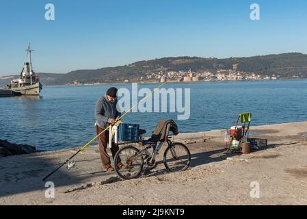 A Turkish fisherman on the pier at Canakkale, with the Dardanelles Strait, Gallipoli Peninsula and Kilitbaher in the background. The Gallipoli Peninsula on the north bank of the Dardanelles strait in north-western Turkey, is the site of extensive First World War battlefields and memorials, a campaign that took place between the Ottoman Empire and the Allied powers. The Dardanelles Strait is an important commercial shipping route that connects the Mediterranean and the Black Sea, with the city of Canakkale at the entrance, a city that is not only important for its historical and mythological hi Stock Photo