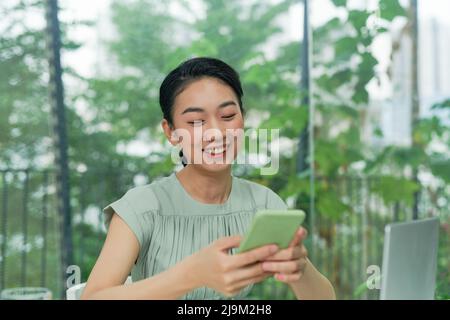 Happy young elegant businesswoman scrolling in smartphone while enjoying break by workplace Stock Photo
