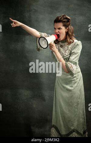 Vintage portrait of young shouting girl in medieval style dress with megaphone isolated on dark background. Comparison of eras concept. News, sales Stock Photo