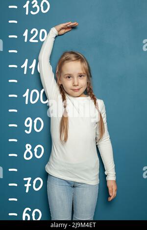 Cute little girl measuring height near blue wall background at home Stock Photo